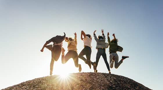 Rear view of group of happy friends having fun on mountain top. Men and women jumping on mountain top against sunset.