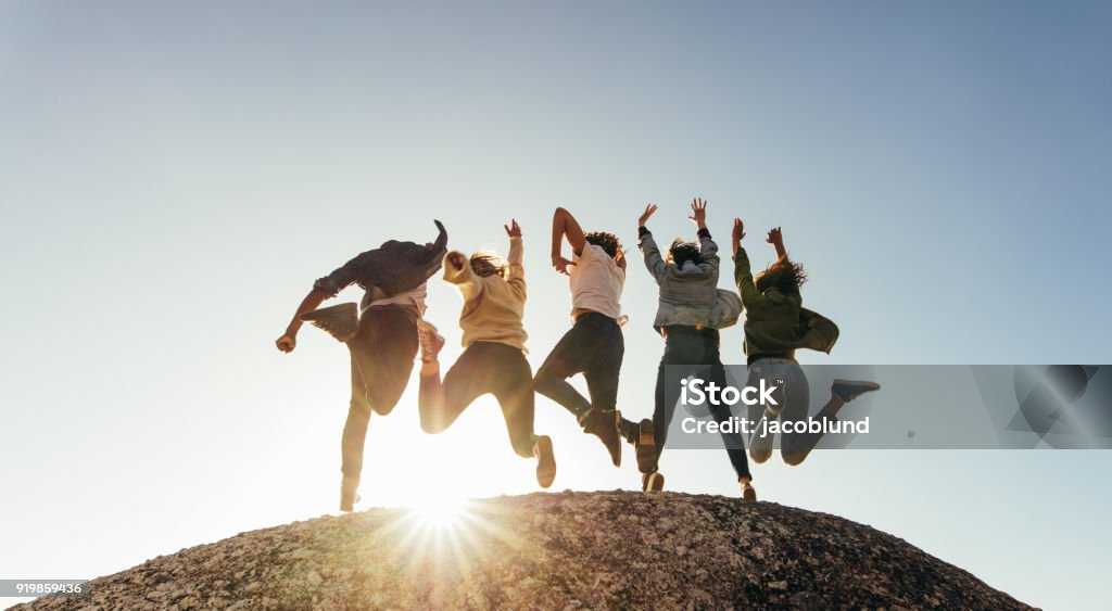 Group of happy friends having fun on mountain top Rear view of group of happy friends having fun on mountain top. Men and women jumping on mountain top against sunset. Jumping Stock Photo