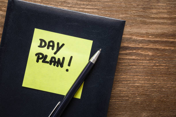 istock Inscription day plan written on yellow sticky note. Notebook planner with pen on the brown wooden office desk. Schedule concept. 919912442