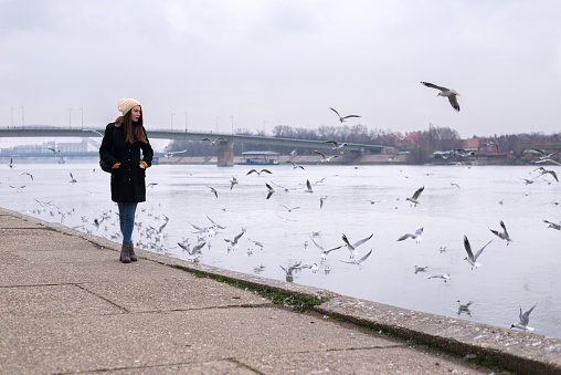 Young woman walking on the pedestrian walkway beside the river in the city and watching seagulls flying.