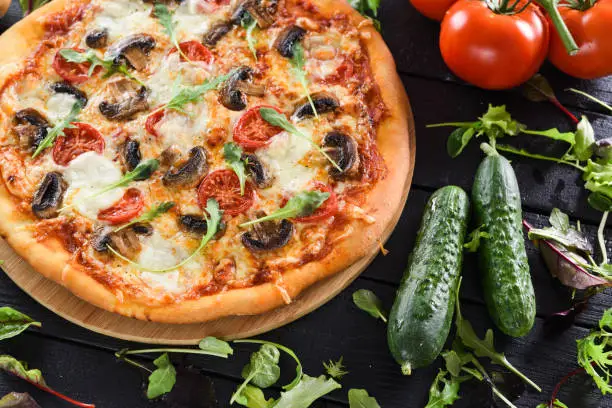Delicious homemade pizza with mushrooms, tomatoes, mozarella and arugula served with fresh vegetables and herbs on black background closeup