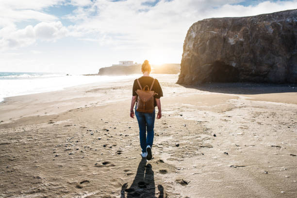 istock back view of young blonde woman with leather backpack walking on empty beach at sunset 919986774