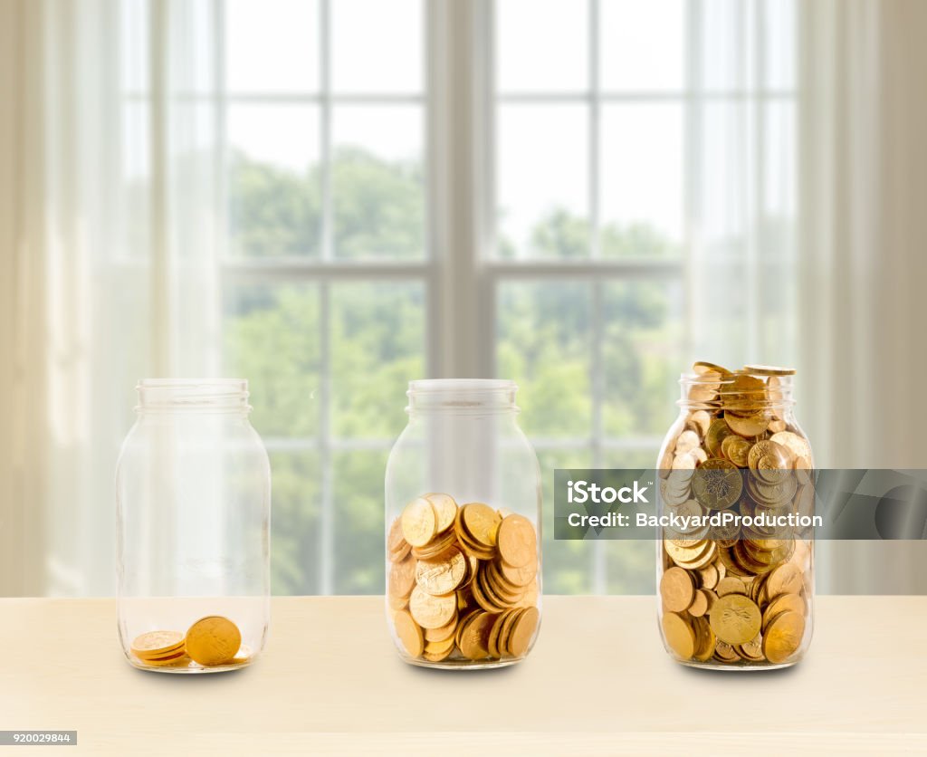 Three glass jars of growing savings on shelf Concept of investment or savings for retirement with three glass jars filled with gold coins in front of bright window Coin Stock Photo