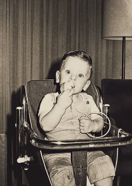 Vintage black and white grainy image of a cute kid on a baby chair eating.