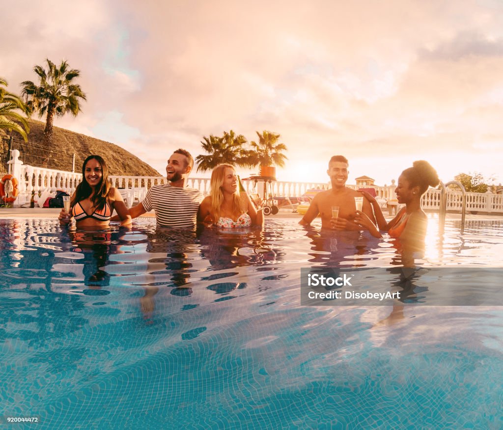 Happy friends drinking champagne in pool party at sunset - Rich people having fun in exclusive tropical vacation - Holiday, youth lifestyle and friendship concept - Main focus on left guys Tourist Resort Stock Photo