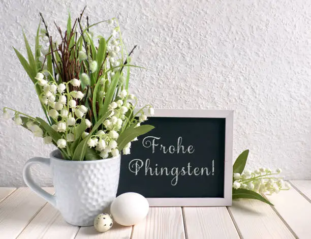 Easter composition with blackboard decorated with ceramic hen, eggs and and lily of the valley flowers. Caption "Frohe Phingsten" means "Happy Pentecost" in German.