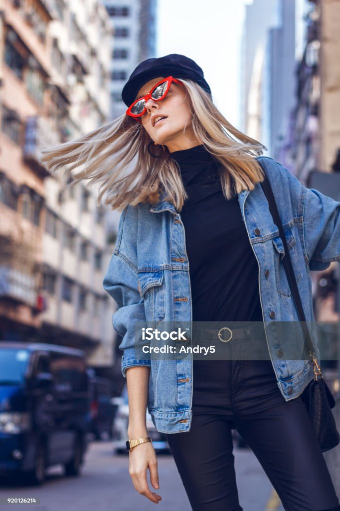 Beautiful woman with red sunglasses A beautiful young blond woman with fashionable red sunglasses walking through a small urban street in Hong Kong. She is dressed in black clothes with a denim jacket and dancing on the street. Fashion Stock Photo