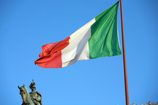 Picture of the Italian flag on a pole blowing in the wind with a statue of Victor Emmanuel II on a horse behind it, both under a clear, blue sky. The flag has three vertical stripes in green, white, and red. The Vittorio Emanuele II statue, designed by Giuseppe Sacconi at the end of 19th century, is a monument to honor Victor Emmanuel II, the first king of a unified Italy. 