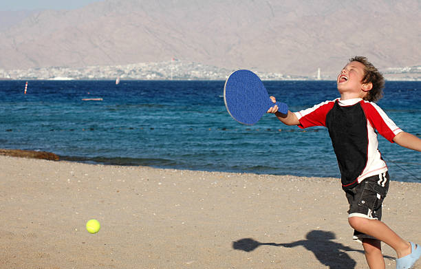 happiness on the beach A boy hitting a ball with a racket on the shores of the Red Sea, Eilat, Israel (Aqaba, Jordan is in the background) neta degany stock pictures, royalty-free photos & images