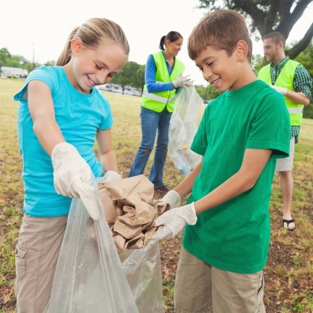 African American boy and girl wear gloves picking up plastic bottles into a black garbage bag outdoor in the park. Two children picking garbages outside. Volunteer and charity concept