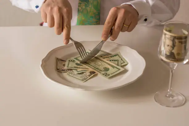 Money on a plate being cut like food with a knife and fork by two human hands