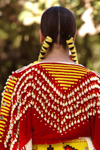 A young American Indian girl in colorful traditional costume with braided hair at a pow wow in Hawaii.  Ribbons and shells adorn her braided hair and patterned clothing. A vertical close-up color photograph of the back of a young girl, selective focus, narrow depth of field, with red, yellow, white and green colors, shot outdoors under natural light conditions.