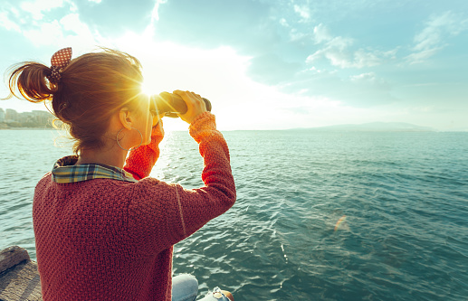 Young Girl Looking Through Binoculars At The Sea On A Bright Sunny Day, Rear View. Wanderlust Travel Journey Concept