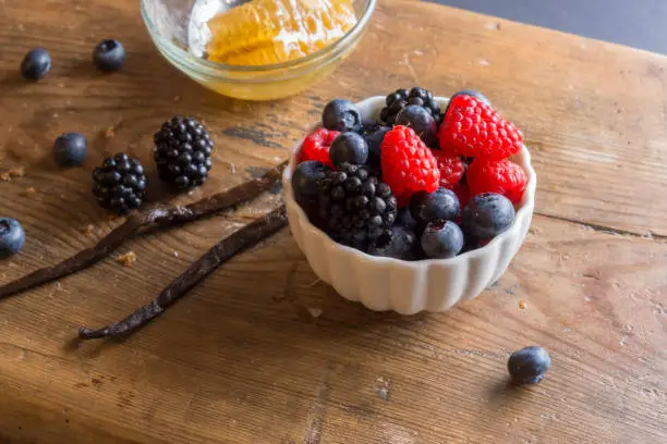 Blackberries, Raspberries, Bilberries in a white bowl, Vanilla Bean and Honeycomb with a spoon on a wooden surface