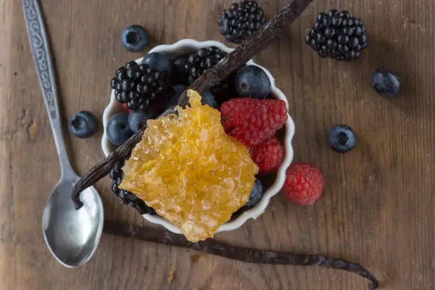 Blackberries, Raspberries, Bilberries in a white bowl, Vanilla Bean and Honeycomb with a spoon on a wooden surface