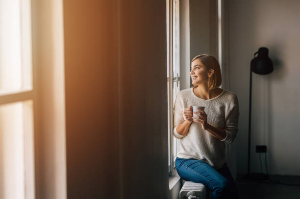 istock Beautiful young woman drinking coffee and looking through window while sitting at windowsill at home. 920406312