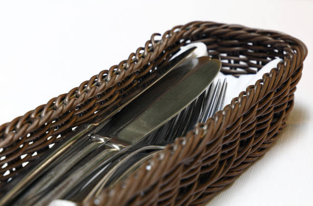 Close up metal dining utensils, forks and knives, in wooden basket container on white tablecloth, high angle view, personal perspective