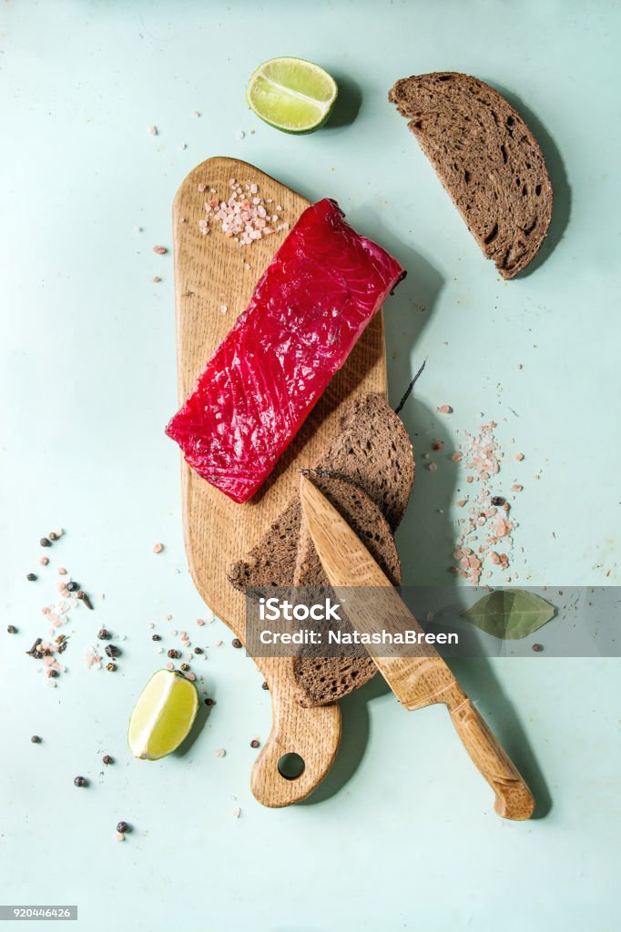 Beetroot marinated salmon Piece of beetroot marinated salted salmon with sliced rye bread, pink salt, pepper and lime served on wooden cutting board over light green pin-up style background. Top view, space Above Stock Photo