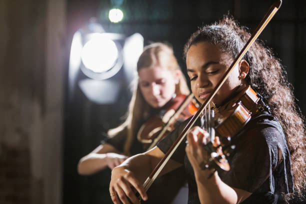 Teenage girls playing violin in concert Two teenage girls playing the violin on stage. The focus is on the 15 year old mixed race girl in the foreground. She has a serious expression on her face, concentrating and looking down as she plays her instrument. stage performance space stock pictures, royalty-free photos & images