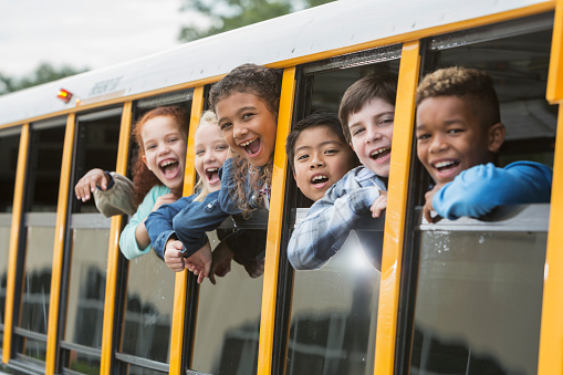 A group of six multi-ethnic elementary school students, 7 to 9 years old, on a yellow school bus, looking out the windows. The view is from outside the bus looking in.