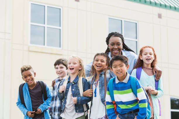 Teacher and multi-ethnic children carrying backpacks A mature African-American woman in her 40s, the teacher, standing outside a school building with a group of multi-ethnic school children wearing casual clothing and carrying backpacks. They are looking at the camera, smiling and laughing. elementary student with teacher stock pictures, royalty-free photos & images