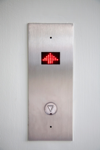 A very basic image of a elevator floor sign with the arrow pointing up. The wall is grey and the elevator sign plate is silver. The arrow is red and small. Also there is only one button to call the elevator to go down so, the image was taken on the top floor. 