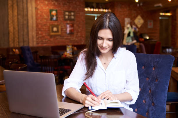 istock Swarthy beautiful girl is planning her week in the diary. The brunette creates problems and solutions to them. A young woman sits in a restaurant, on the table is a laptop for work. 920503624