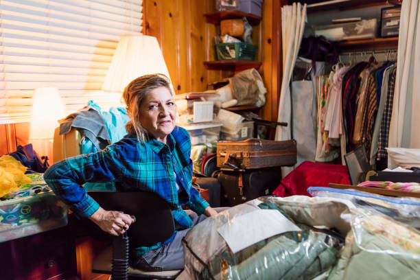 Senior woman at home, messy room A senior woman in her 60s at home, sitting in a messy, cluttered room, looking at the camera with a serious expression. greed stock pictures, royalty-free photos & images