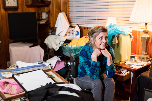 A senior woman in her 60s at home, sitting in a messy, cluttered room, looking away with a serious expression.