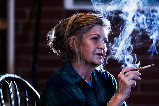 A senior woman in her 60s smoking a cigarette in a dark room. She is looking away with a serious expression.