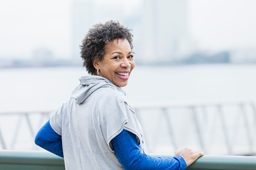 A mature African-American woman in her 50s standing outdoors wearing a hooded shirt on a city waterfront, looking over her shoulder smiling at the camera.