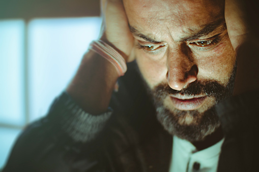 Indoor image of disturbed, sad, Asian, Indian mid adult man with strong character and facial hair. He is sitting at home near door in day time. He is looking down and holding his head while thinking something deeply with blank expression.