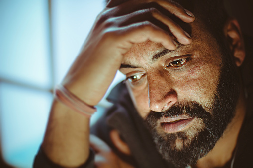 Indoor close-up image of disturbed, sad, Asian, Indian mid adult man with strong character and facial hair. He is sitting at home near door in day time. He is crying and a drop of tear coming out of his eye. He is looking down and holding his head while thinking something deeply with blank expression.