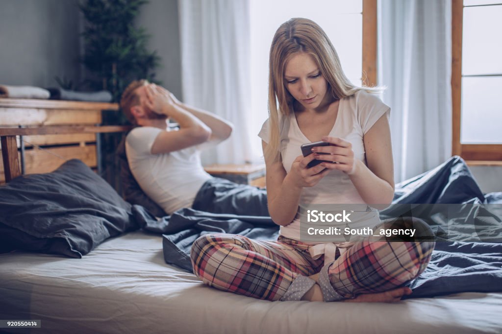 Dishonesty Young man having problem while his girlfriend using smartphone in bed Infidelity Stock Photo