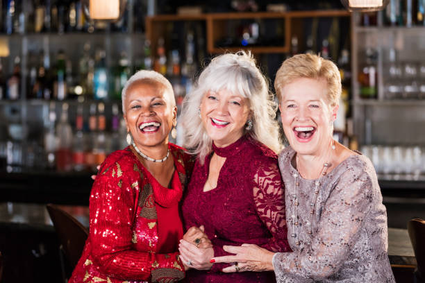 Three multi-ethnic senior women at a bar A group of three multi-ethnic senior women wearing cocktail dresses, enjoying ladies' night out. They are standing together at a restaurant bar, laughing, looking toward the camera.  The woman on the right is in her 70s and her friends are in their 60s. cocktail dress stock pictures, royalty-free photos & images