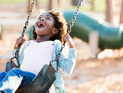 A 5 year old African-American girl playing on a playground, having fun swinging on a swing.