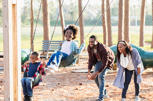 An African-American family playing at a park together, having fun. The children, a 7 year old boy and 5 year old girl are on a swingset and their parents are pushing their swings.