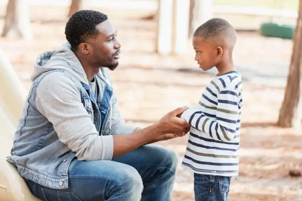 An African-American man in his 30s with a serious expression on his face, talking to his 7 year old son on a playground. They are face to face, and he is holding his hands. The boy may have been naughty and is being disciplined.