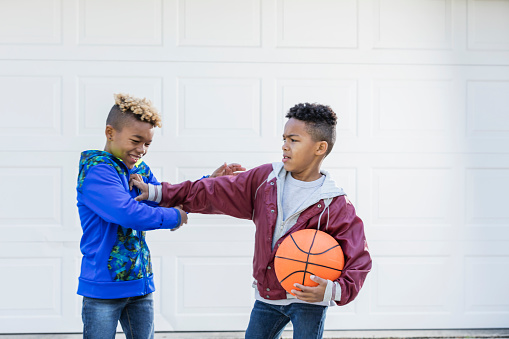 Two mixed race African-American and Caucasian boys, brothers 8 and 10 years old, outdoors on their driveway, playing basketball. The younger one is holding the ball under his arm, pushing his sibling with the other.