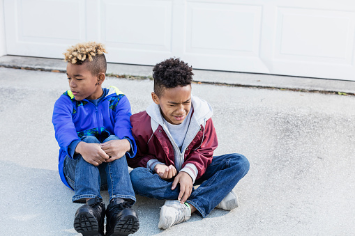 Two mixed race African-American and Caucasian boys, brothers 8 and 10 years old, outdoors sitting on their driveway. The younger one is crying or frowning and his sibling is looking away, ignoring him.