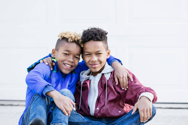 Two mixed race African-American and Caucasian boys, brothers 8 and 10 years old, outdoors sitting on their driveway side by side, arms around each other's  shoulders, smiling at the camera.