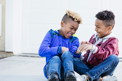 Two mixed race African-American and Caucasian boys, brothers 8 and 10 years old, outdoors sitting on their driveway side by side, laughing together.