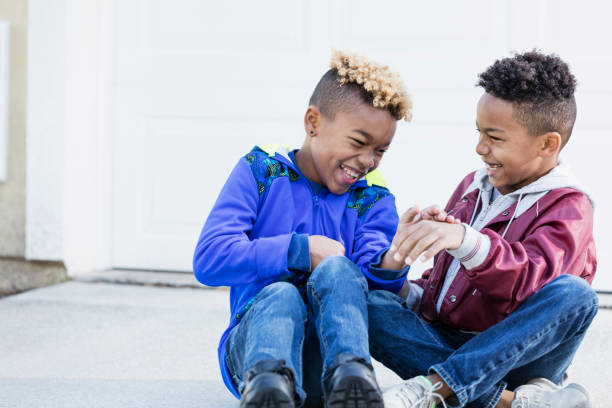 Little boys, brothers and best friends, laughing Two mixed race African-American and Caucasian boys, brothers 8 and 10 years old, outdoors sitting on their driveway side by side, laughing together. family with two children stock pictures, royalty-free photos & images