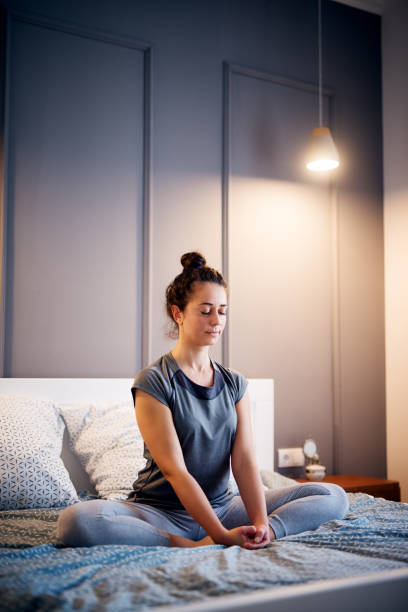 Peaceful pretty middle aged active woman practising yoga on the bed holding eyes closed, sitting in Half Lotus exercise, Ardha Padmasana pose and wearing sportswear at night.