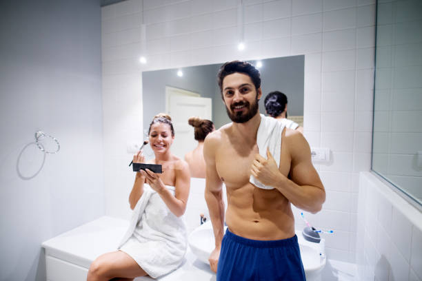 istock Young muscular shirtless man holding a towel on the shoulder while looking at the camera and his girlfriend making make-up with a brush in the morning at the bathroom. 920828556