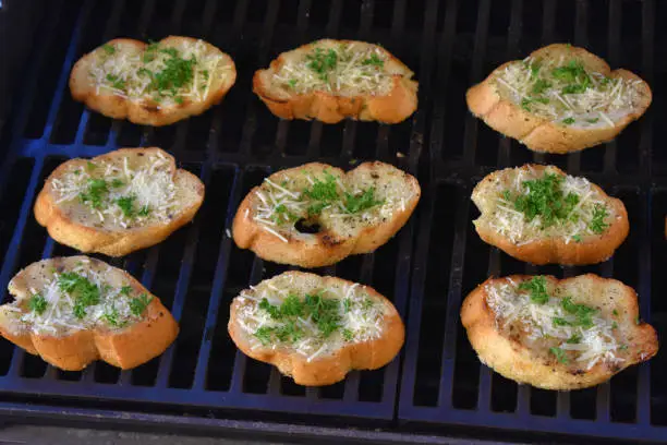 Slices of garlic bread toasting on the grill and topped with parmesan cheese and chopped parsley