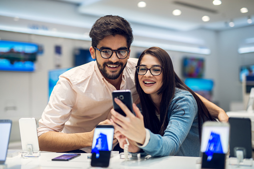 Close up focus view of a stylish charming embracing young student love couple taking a selfie with new mobile in a tech store.