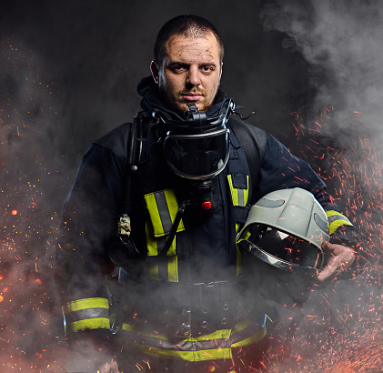 A professional firefighter dressed in uniform holding safety helmet in fire sparks and smoke over a dark background.