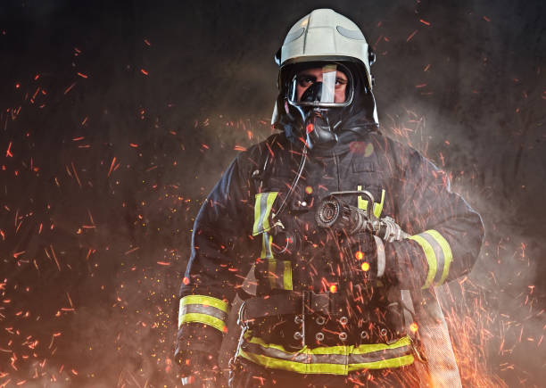 A firefighter dressed in a uniform in a studio. A professional firefighter dressed in uniform and an oxygen mask standing in fire sparks and smoke over a dark background. extinguishing photos stock pictures, royalty-free photos & images