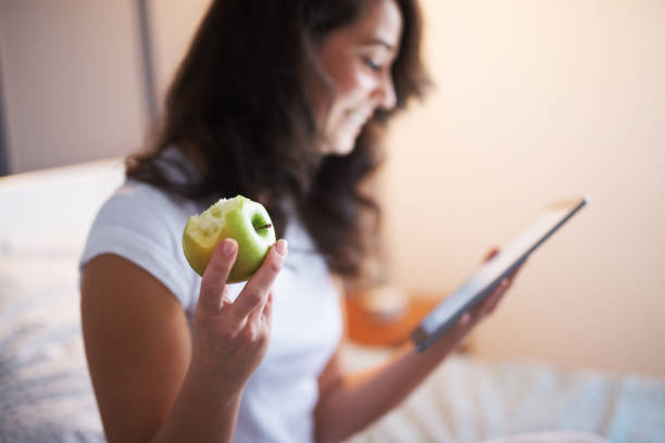 Close up of smiling beautiful middle aged woman sitting on the bed and holding an apple while looking in a tablet. Hand and bitted apple in focus. Close up of smiling beautiful middle aged woman sitting on the bed and holding an apple while looking in a tablet. Hand and bitted apple in focus. apple with bite out of it stock pictures, royalty-free photos & images